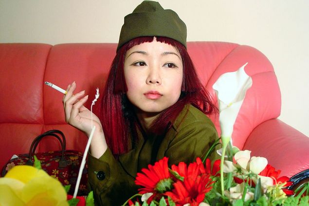 Video still of a young Asian woman in khaki uniform, holding a cigarette, with a bouquet of flowers in front of her, which makes up the lower frame. 