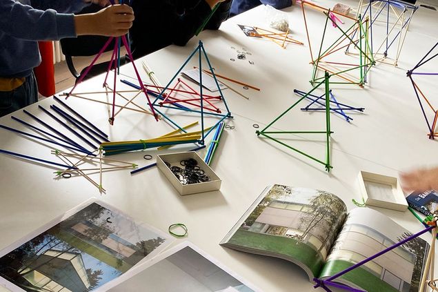 On a table are architectural pictures from the Goetz Collection, next to which children build spatial structures out of colorful straws