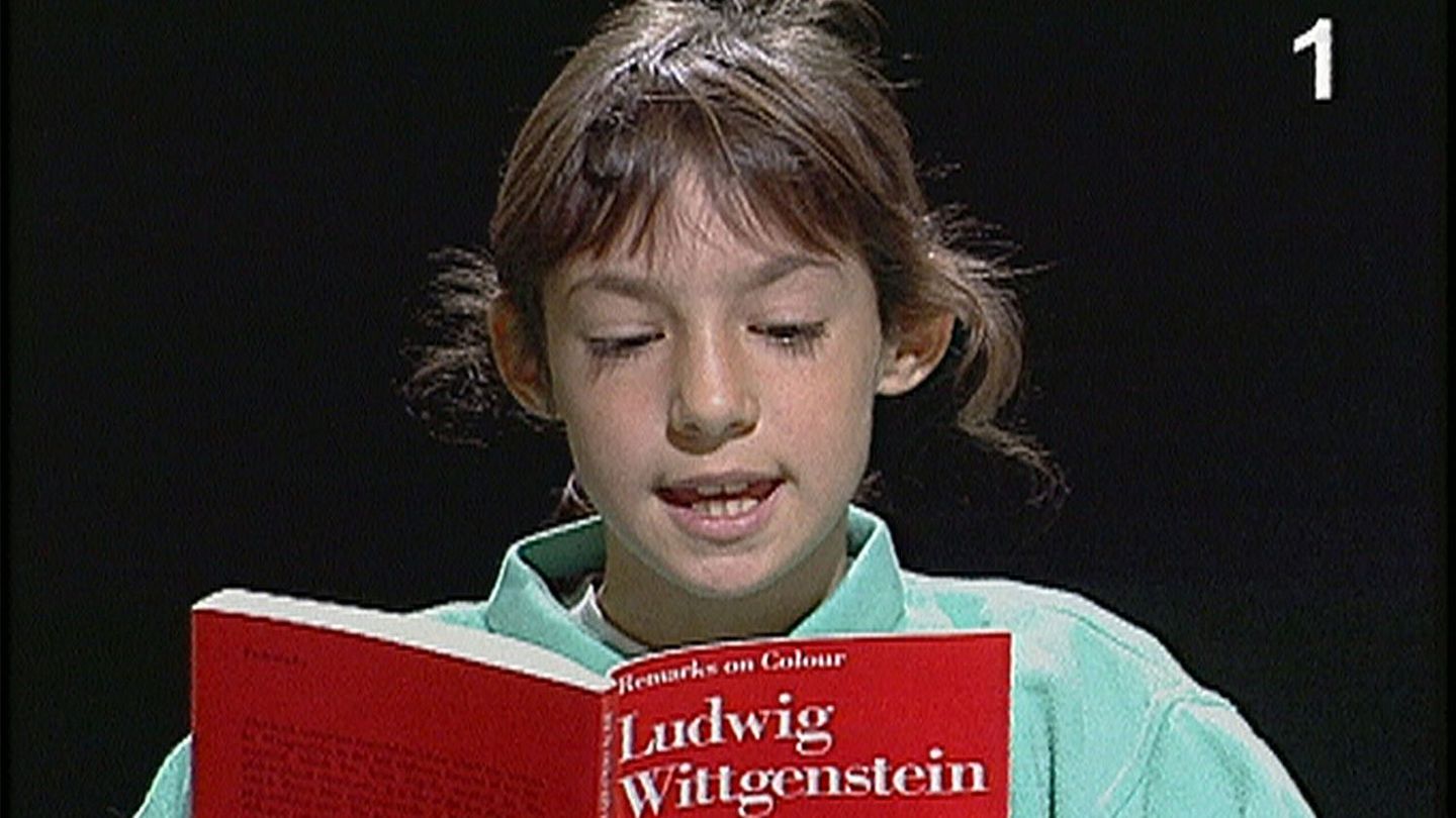 This video still shows a girl of about ten years of age with light brown hair reading from a red edition of the book "Bemerkungen über die Farben" by Ludwig Wittgenstein. 